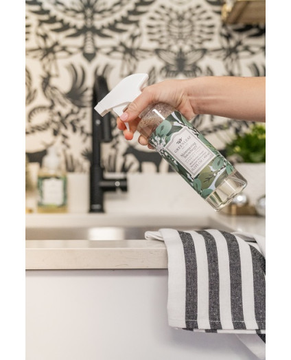 En hand håller Greenleaf Table Top Cleaner med doften Shimmering Snowberries över en köksbänk med svartvit mönstrad tapet, randig handduk och dekorativa detaljer – fräsch och stilren rengöring från House of Månsson