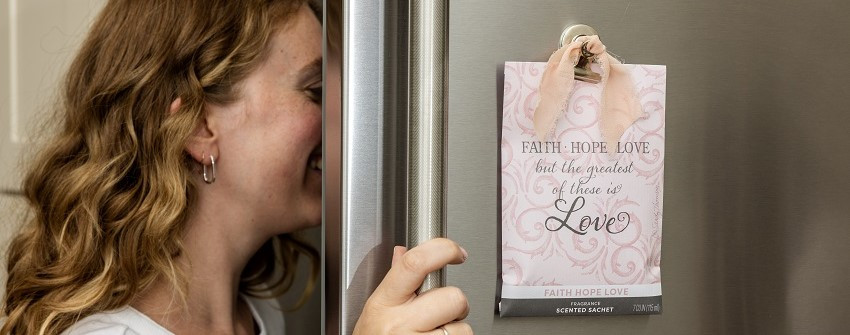 Doftpåse Fresh Scents Faith Hope Love i rosa design med texten "but the greatest of these is Love", hängande på en kylskåpsdörr med sidenband – House Of Månsson