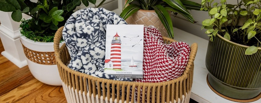 Doftpåse Fresh Scents Tradewinds med motiv av rödvit fyr och hav, placerad i en korg med mönstrade filtar bredvid gröna krukväxter – House Of Månsson
