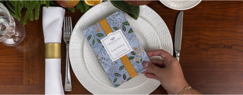 Greenleaf Laurel Grove doftpåse placerad på vit tallrik i elegant bordsdukning med bestick, servett och dekorativa gröna inslag. Exklusiv hemdoft från House Of Månsson.